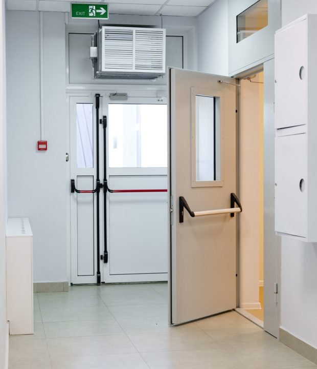 A well-lit corridor features white walls, a closed cabinet, and an open door leading to an exit. The space appears clean and organized, suggesting a functional environment.